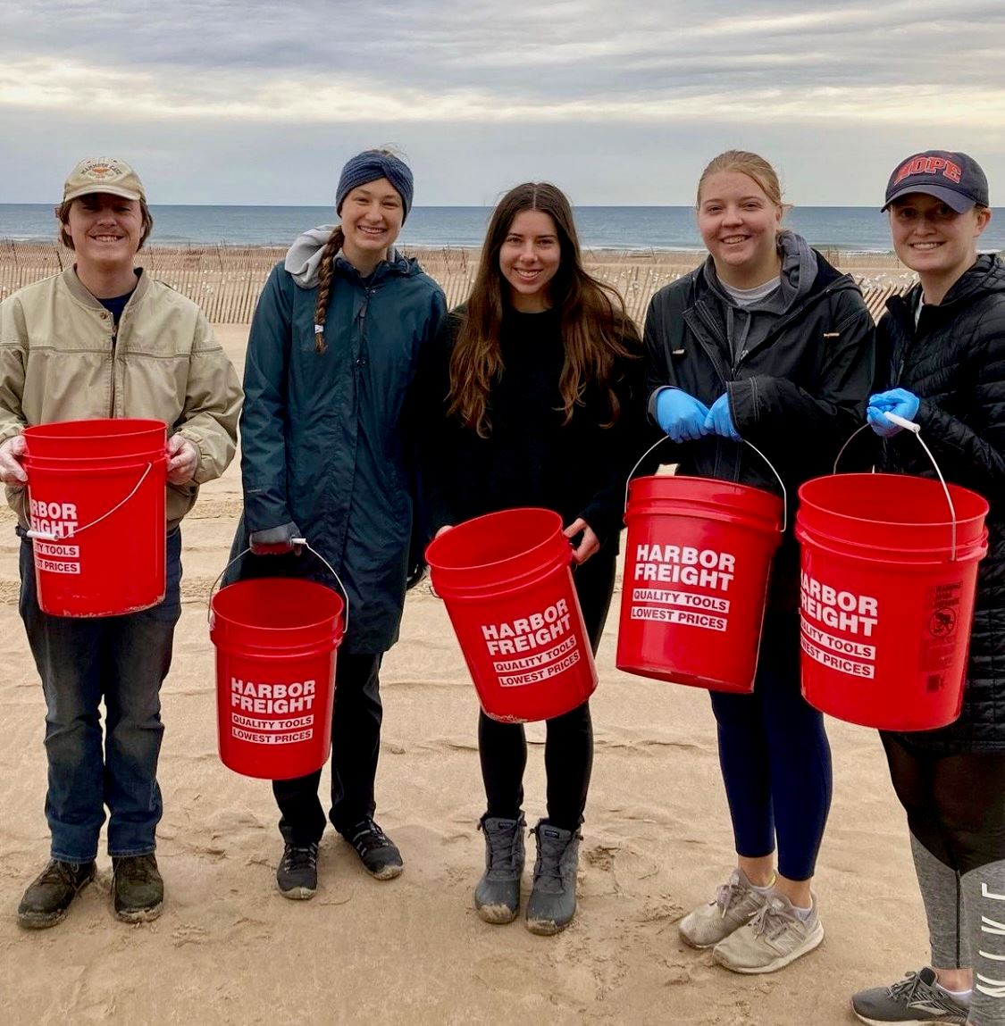 5 college students standing on the beach hold large red buckets for a beach clean up