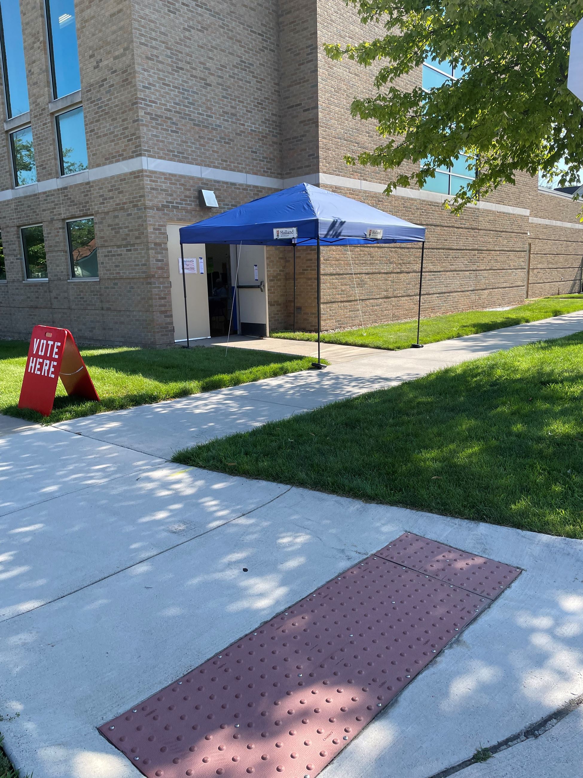 blue canopy tent at a side entrance of a brick building with a sidewalk and grass along the side