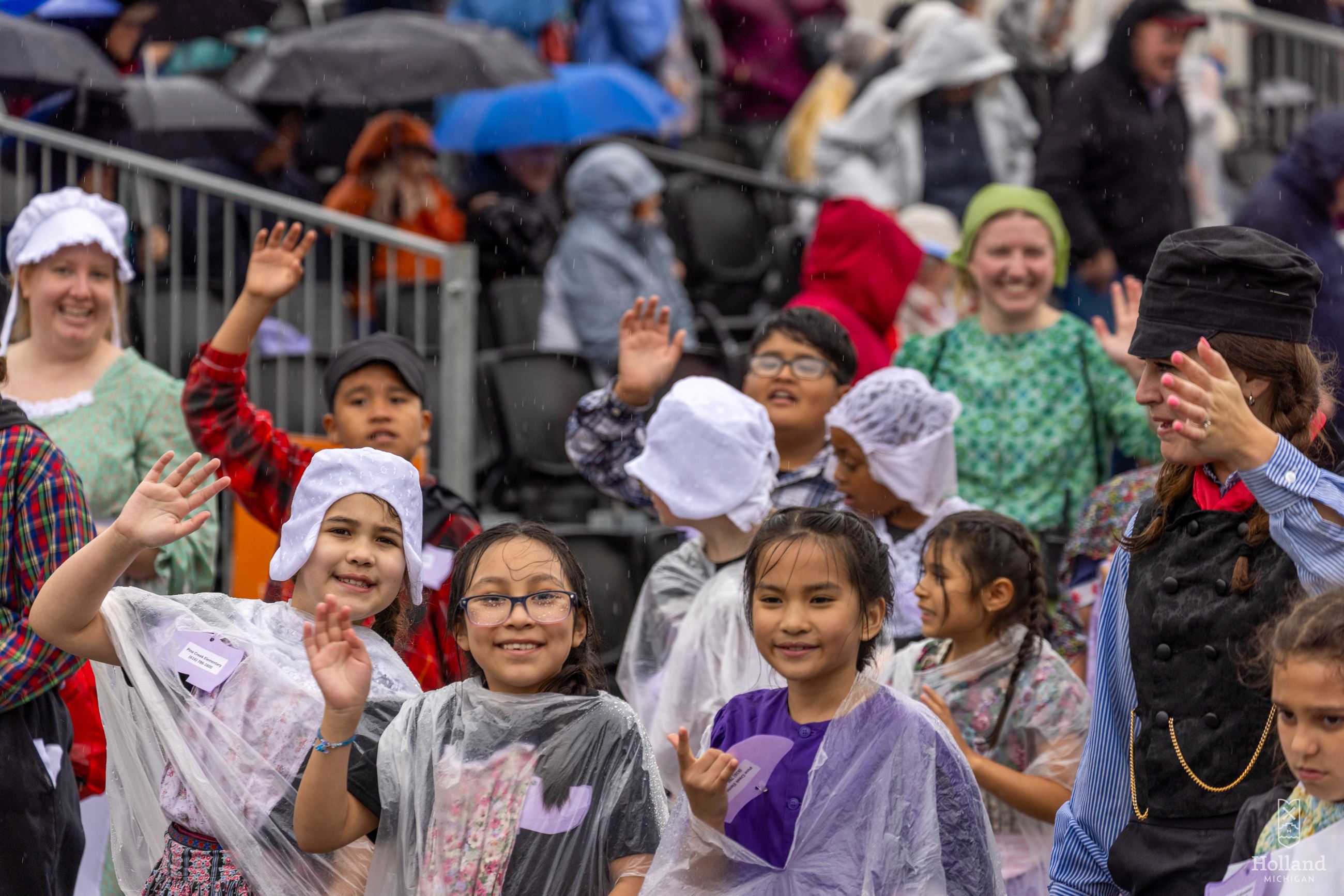 3 young girls dressed in Dutch costumes walking in a parade, waving at the camera