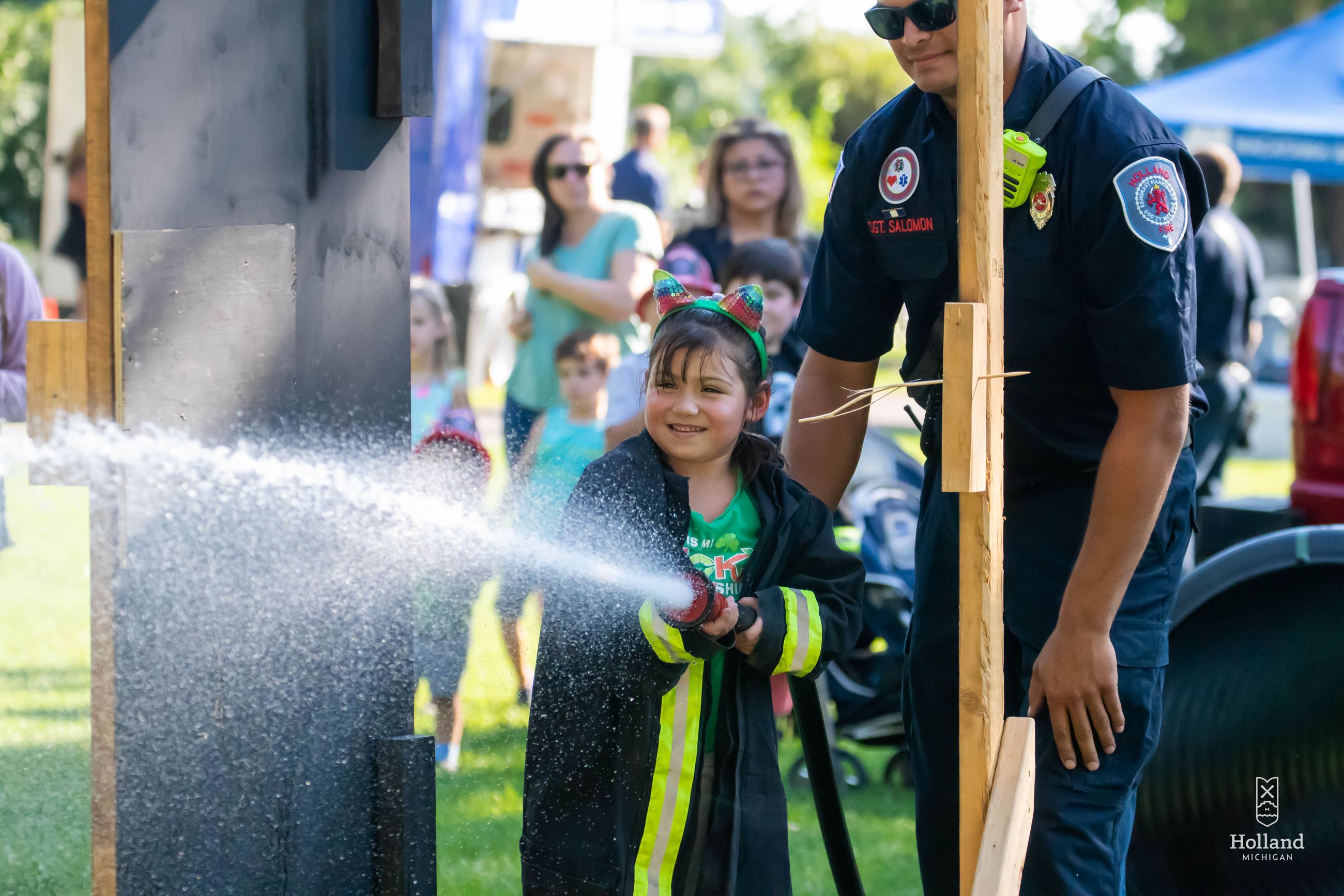 Young girl smiling in an oversized firefighter's jacket holding a fire hose, firefighter standing