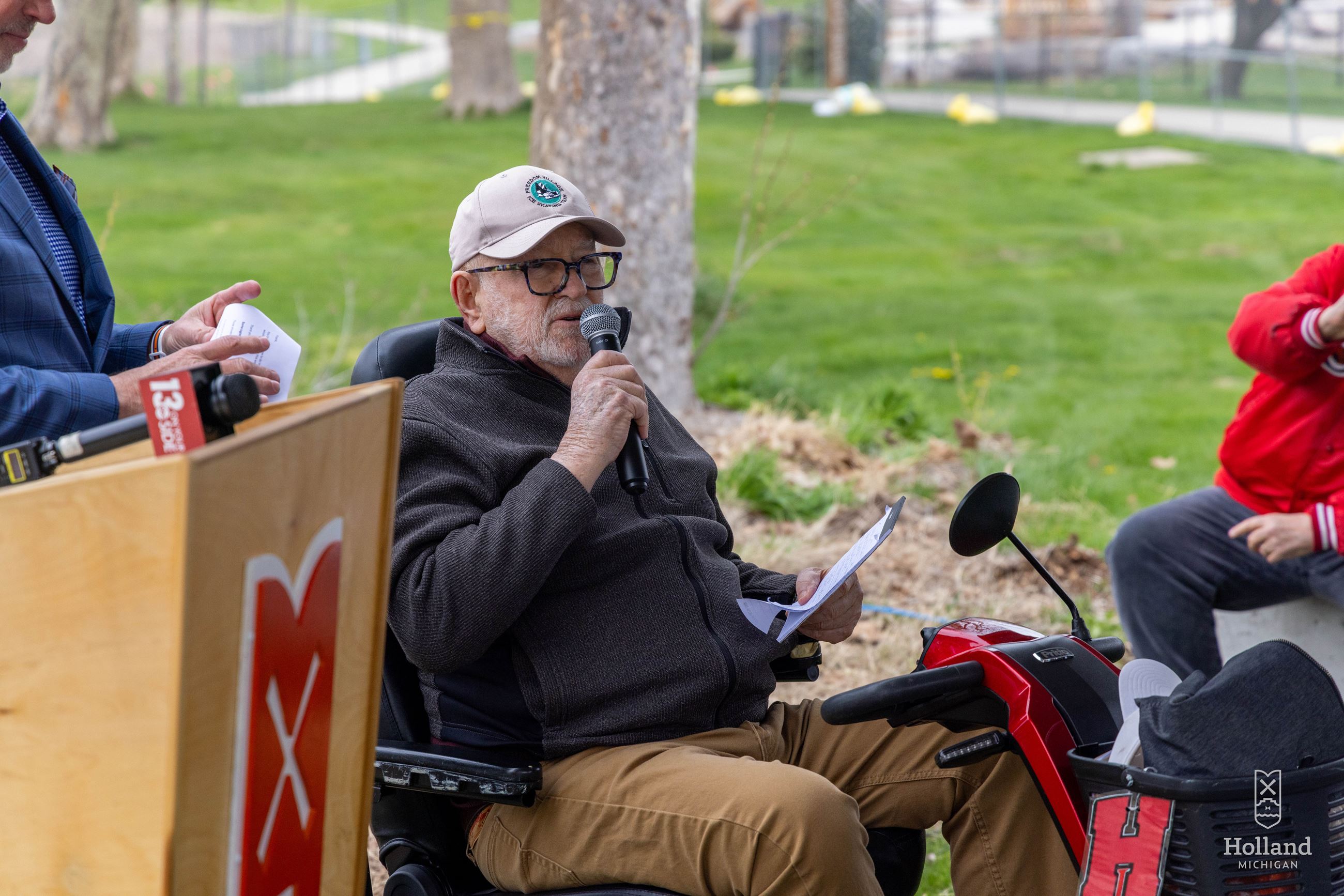 Elderly man in motorized scooter, next to podium, outside speaking with microphone in hand